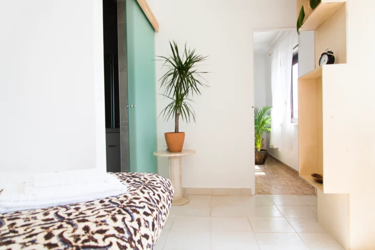 A bright, minimal bedroom in Ericeira with a bed covered in a leopard print blanket, a potted plant on a small round table, and sunlight streaming in from an adjacent room with another plant visible—perfect for peaceful yoga mornings.