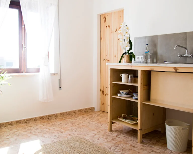 A bright, minimalist kitchen in an Ericeira guest house features a wooden cabinet, open shelves with dishes, a sink, an orchid plant, food prep space, a window with sheer curtains, a closed door, and a small trash bin on tiled floors.