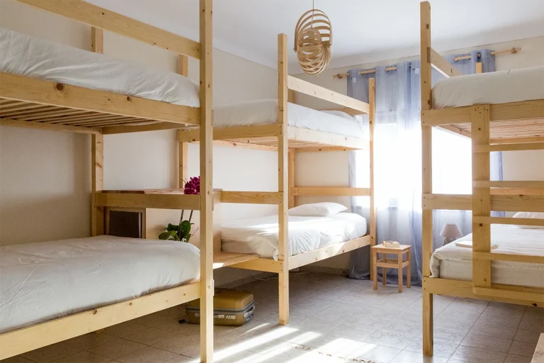 A bright guest house room in Ericeira with four wooden bunk beds and white bedding. Sunlight streams through sheer curtains, highlighting a small stool by the window and a potted plant, perfect for post-yoga relaxation.