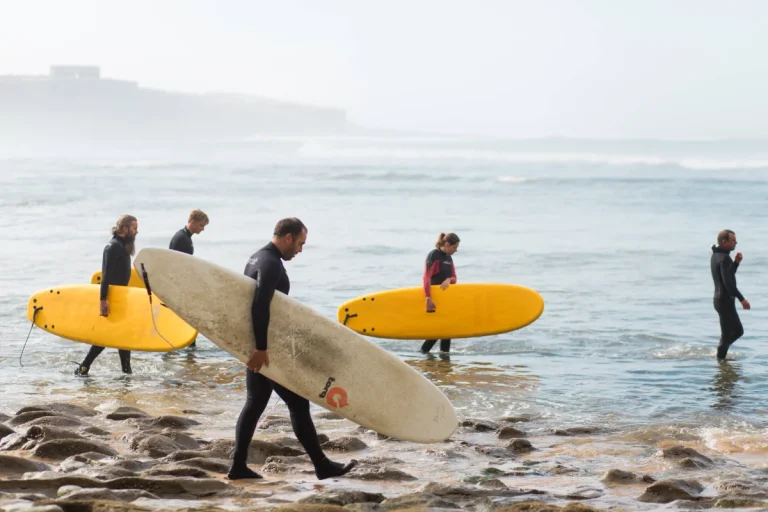 Five surfers in wetsuits carry mostly yellow surfboards as they walk on rocky sand toward the ocean. The hazy sky and distant waves set the scene near a cozy guest house known for its delicious vegan food.