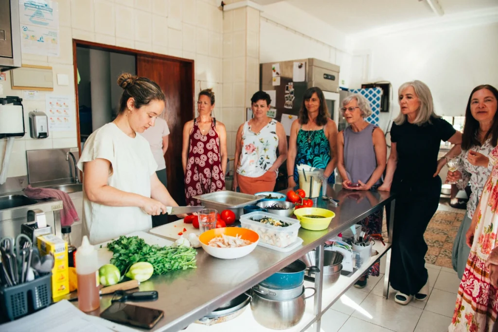 A woman chops vegetables at a kitchen counter while a group of women watches her, enjoying the relaxed vibe. Bowls and trays filled with fresh food create a bright, casual scene—perfect for a guest house gathering after yoga.