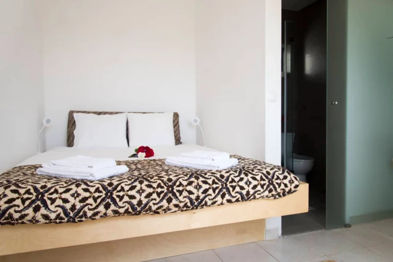 A neatly made bed with a patterned brown and cream blanket, two stacks of folded white towels, and a small bouquet of flowers create a serene setting. This minimalistic Ericeira room with white walls and an open bathroom door is perfect for yoga retreats.