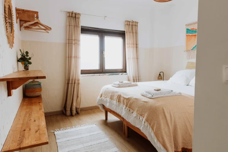 A cozy bedroom in Ericeira with a double bed, beige bedding, two folded towels, wooden shelves, a window with beige curtains, and a woven basket. The room features neutral decor, natural light, and is perfect for enjoying local vegan food nearby.