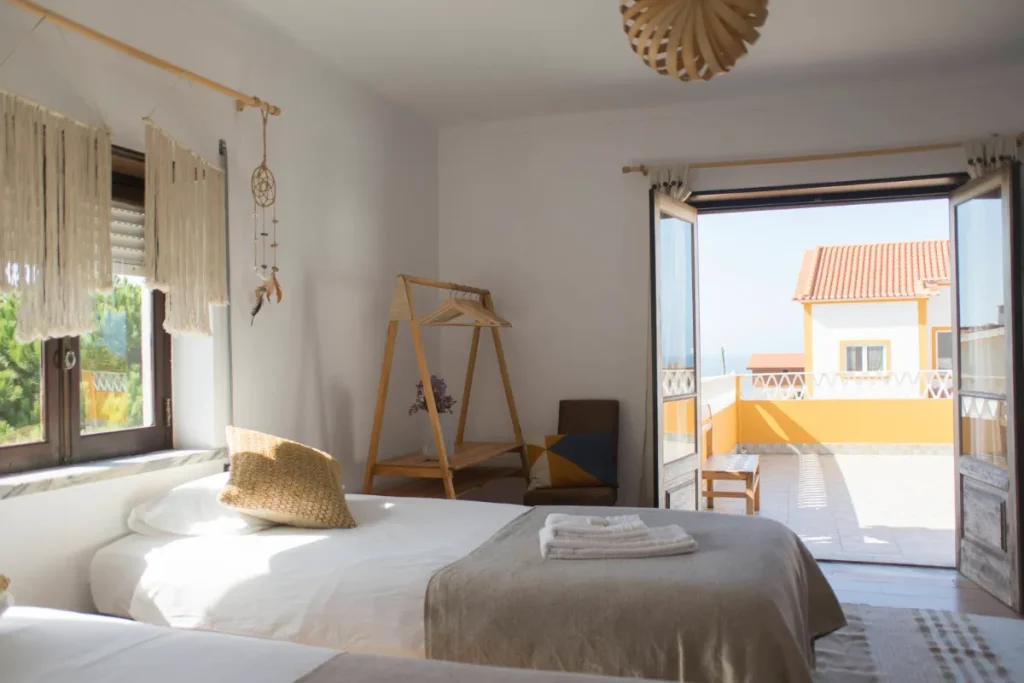 A bright, cozy bedroom in an Ericeira guest house with two single beds, a window, a wooden clothes rack, and open double doors leading to a sunny balcony with views of the sea and nearby buildings.