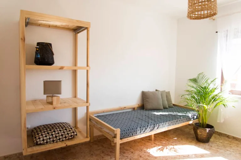 Minimalist guest house room with a wooden daybed, gray patterned bedding, two gray cushions, a wooden shelving unit with a lamp, basket, and cushion, plus a potted plant by the bed and sunlight streaming through the window. Vegan-friendly ambiance throughout.