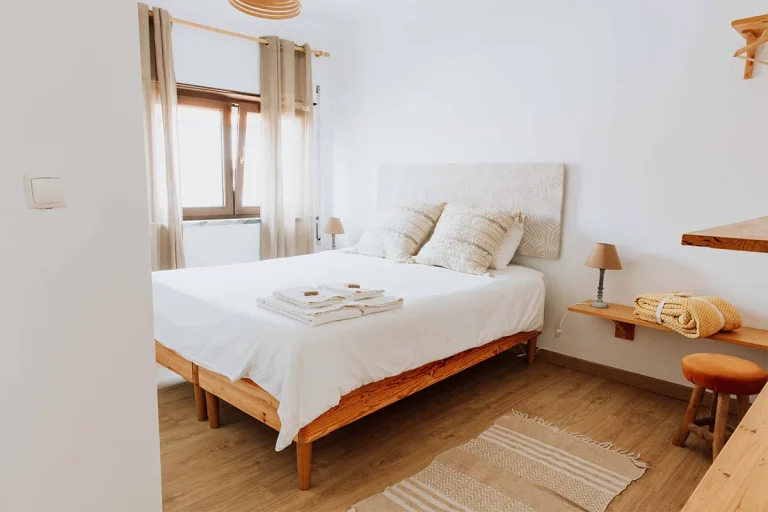 A bright, cozy bedroom at a vegan guest house in Ericeira features a double bed with white bedding, two pillows, and neatly folded towels. Wooden furniture, beige curtains framing a sunlit window, and a rug complete the inviting space.