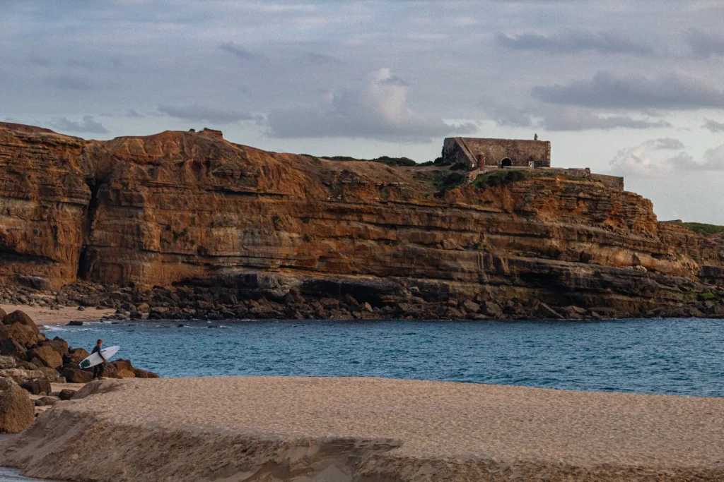 A surfer carrying a board walks along Ericeira's sandy shore near rocky cliffs, with a small guest house perched on top. The ocean and cloudy sky are in the background.