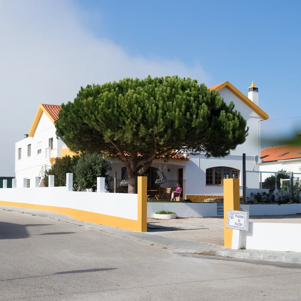 Ondina, a charming white guesthouse with a red-tiled roof stands in Ericeira, shaded by a large pine tree. A person relaxes on the porch, while a white and yellow fence borders the property and a street sign appears in the foreground.