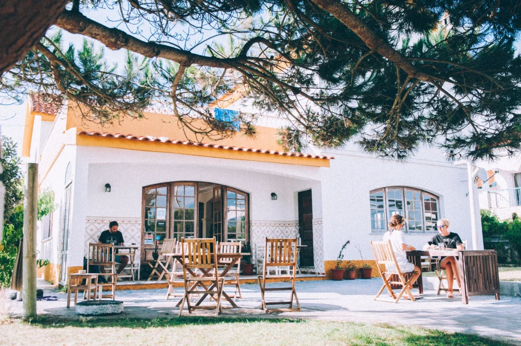 People sit at wooden tables and chairs on the patio of Ondina guesthouse with arched windows, surrounded by trees and greenery in sunny Ericeira.
