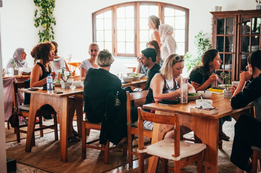 A group of people sit at wooden tables in a bright, cozy café, enjoying vegan food and lively conversation. Sunlight streams through large windows, and plants decorate the space, creating a warm and inviting atmosphere.