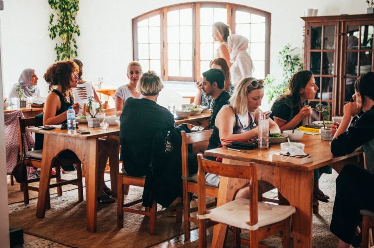 A group of people sit at wooden tables in a bright, cozy café, enjoying vegan food and lively conversation. Sunlight streams through large windows, and plants decorate the space, creating a warm and inviting atmosphere.