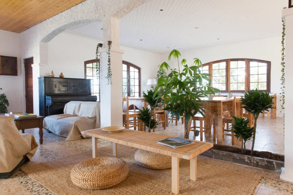 Bright, airy living and dining space with large windows, indoor plants, a piano, wooden furniture, woven poufs, and neutral-colored sofas. Natural light fills the room in true Ericeira style, creating a cozy atmosphere perfect for vegan food gatherings.